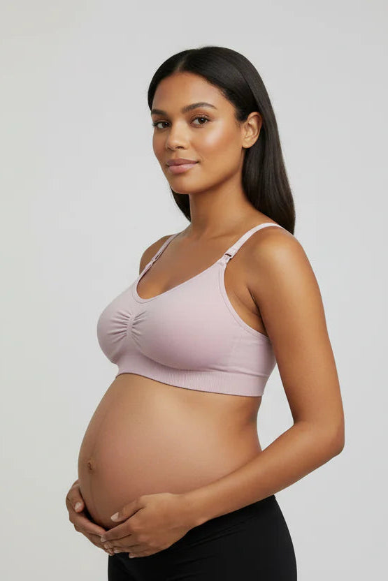 Pregnant woman wearing a light pink maternity bra and black pants on a plain background